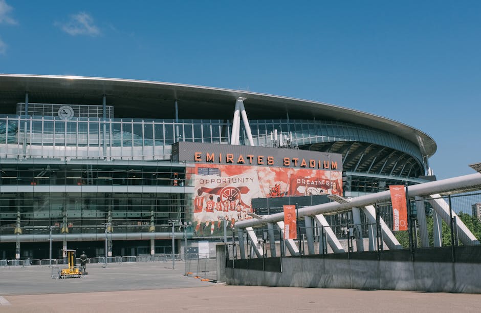Exterior view of the Emirates Stadium in Highbury during daytime, with a clear blue sky overhead. The modern, curved architectural design features a prominent sign reading 'EMIRATES STADIUM' in bold orange letters above multiple glass windows. In the foreground, there are orange banners attached to white poles, and a small area with a black metal fence around the perimeter. The ground is paved, and a man operating a trolley or cart can be seen near the building entrance. This scene depicts a site suitable for home relocation and furniture transport services by Man and Van Highbury, illustrating the logistical aspects of packing and loading objects for moving in an urban environment.