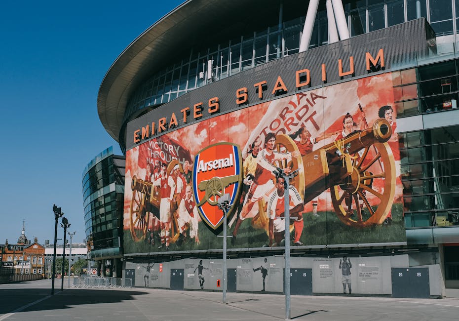 Exterior view of the Emirates Stadium in Highbury during daytime, with a clear blue sky overhead. The modern, curved architectural design features a prominent sign reading 'EMIRATES STADIUM' in bold orange letters above multiple glass windows. In the foreground, there are orange banners attached to white poles, and a small area with a black metal fence around the perimeter. The ground is paved, and a man operating a trolley or cart can be seen near the building entrance. This scene depicts a site suitable for home relocation and furniture transport services by Man and Van Highbury, illustrating the logistical aspects of packing and loading objects for moving in an urban environment.