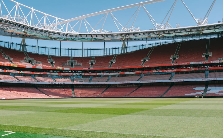 Interior view of a large, modern football stadium with a partially covered roof structure supported by white steel trusses. The seating sections are arranged in multiple tiers and are predominantly red, with some pink and dark green seats visible, lining the curved perimeter of the stadium. The field is well-maintained with lush green grass, featuring clearly marked white lines for gameplay boundaries. The pitch appears to be prepared for a match or event, with construction or maintenance equipment visible on one side near the field’s edge. The sky above the stadium is clear and blue, with natural daylight illuminating the interior. This setting illustrates a spacious venue suitable for sporting events, and reflects the kind of environment where large-scale sports or public gatherings might occur, which could be associated with plans involving logistics for transporting equipment or preparing for a team’s arrival; details of furniture or boxes are not present, but the focus is on the stadium’s structural features and seating arrangement relevant to large-scale event logistics.