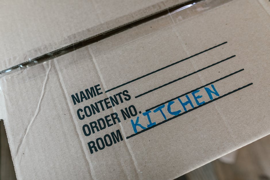 Close-up image of a cardboard moving box, with printed black text labels for 'Name', 'Contents', 'Order No.', and 'Room', where the room label is handwritten in blue ink as 'Kitchen'. The box appears to be sealed with clear packing tape and is positioned indoors, likely on a floor or table, ready for home relocation. The labeling helps facilitate efficient packing and handling during furniture transport and packing processes, which are key aspects of professional removals services like those provided by Man and Van Highbury. The background is blurred, focusing attention on the labeled cardboard box as part of the packing stage for a house move or furniture transport operation.