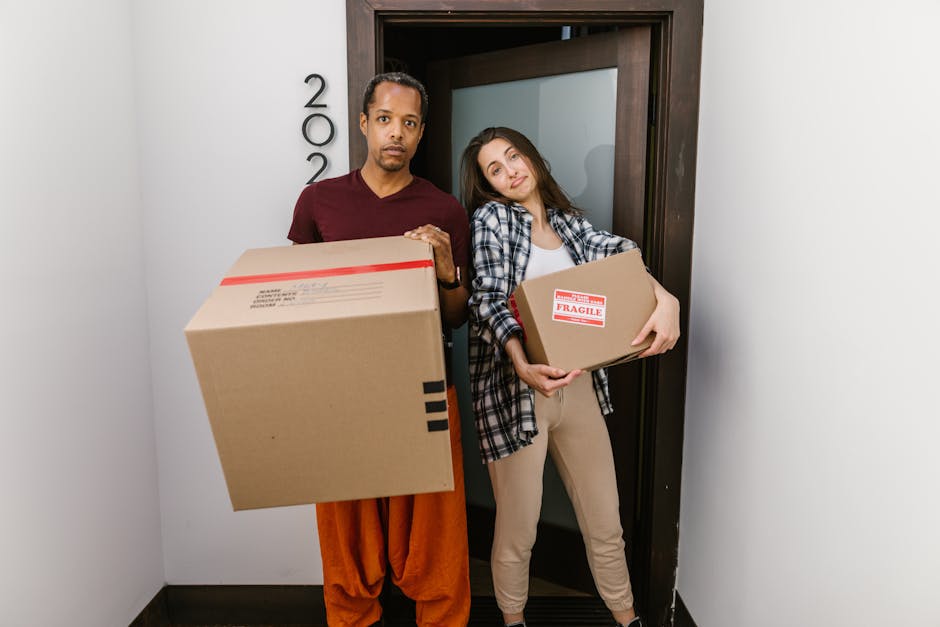 A man and a woman stand inside a doorway, each holding a cardboard box during a home relocation. The man, dressed in a maroon t-shirt and orange trousers, holds a large cardboard box in his right hand, with a slightly serious expression. The woman, wearing a checkered shirt over a white top and beige trousers, carries a smaller cardboard box labeled 'Fragile' in red and white tape, leaning her head slightly towards the man. The background shows the door frame and a white wall with the room number partially visible, and the area appears well-lit, likely with natural light. Visible packing materials include cardboard boxes and plastic straps, and the environment indicates the loading or unloading process associated with furniture transport and packing during removals handled by Man and Van Highbury. This scene captures the logistic steps involved in house moves, emphasizing careful handling of packed items for a professional relocation service.
