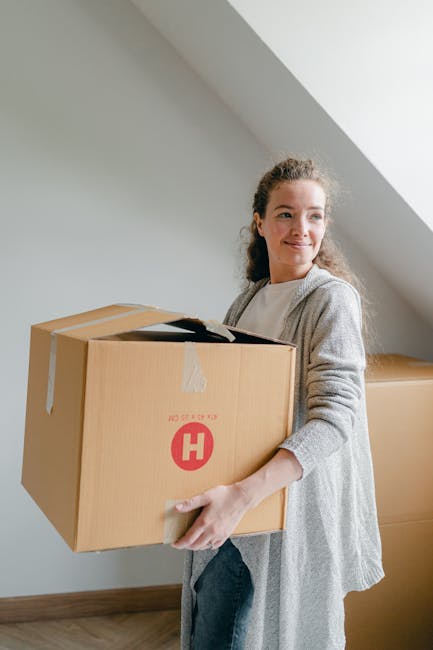 A man wearing a dark grey t-shirt is carrying two large cardboard moving boxes, with a third smaller box balanced on top, on the steps outside a modern residential building. The boxes are secured with