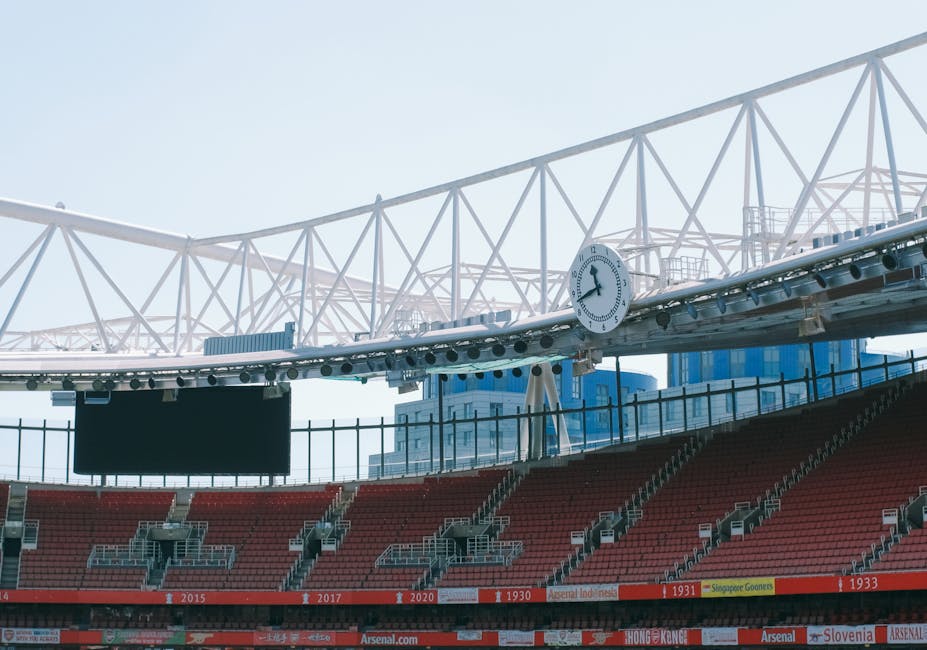 A view inside a sports stadium showing rows of red seating with some metal and plastic barriers and stairways, partially visible in the background is the stadium's exterior structure with a white lattice roof support and a large clock displaying 10:10. The lighting suggests daytime with a clear sky. Although the image primarily captures the stadium environment, it relates to the context of house removals and logistics where equipment, packing materials, and vehicles such as removal vans might be used in the process. Occasionally, Man and Van Highbury's services include loading and transport operations that can involve coordination of such facilities and timing, as indicated by the visible clock and seating arrangement.