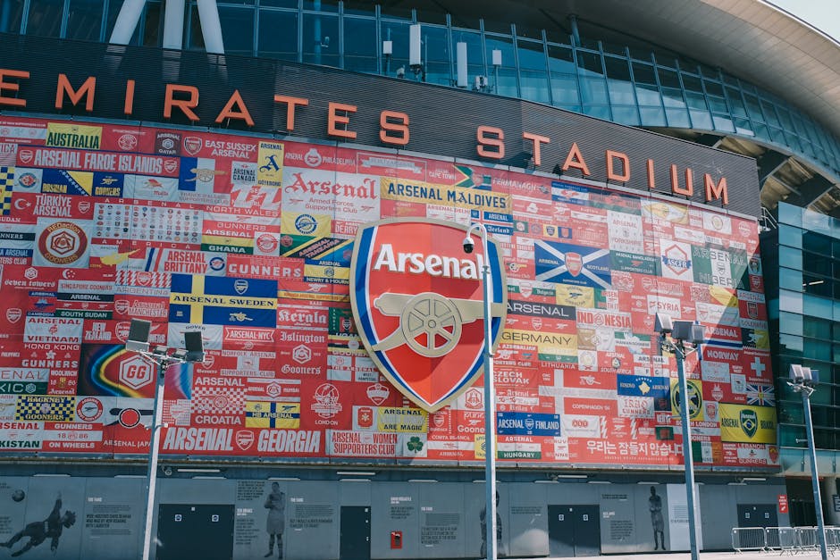 A view inside a sports stadium showing rows of red seating with some metal and plastic barriers and stairways, partially visible in the background is the stadium's exterior structure with a white lattice roof support and a large clock displaying 10:10. The lighting suggests daytime with a clear sky. Although the image primarily captures the stadium environment, it relates to the context of house removals and logistics where equipment, packing materials, and vehicles such as removal vans might be used in the process. Occasionally, Man and Van Highbury's services include loading and transport operations that can involve coordination of such facilities and timing, as indicated by the visible clock and seating arrangement.