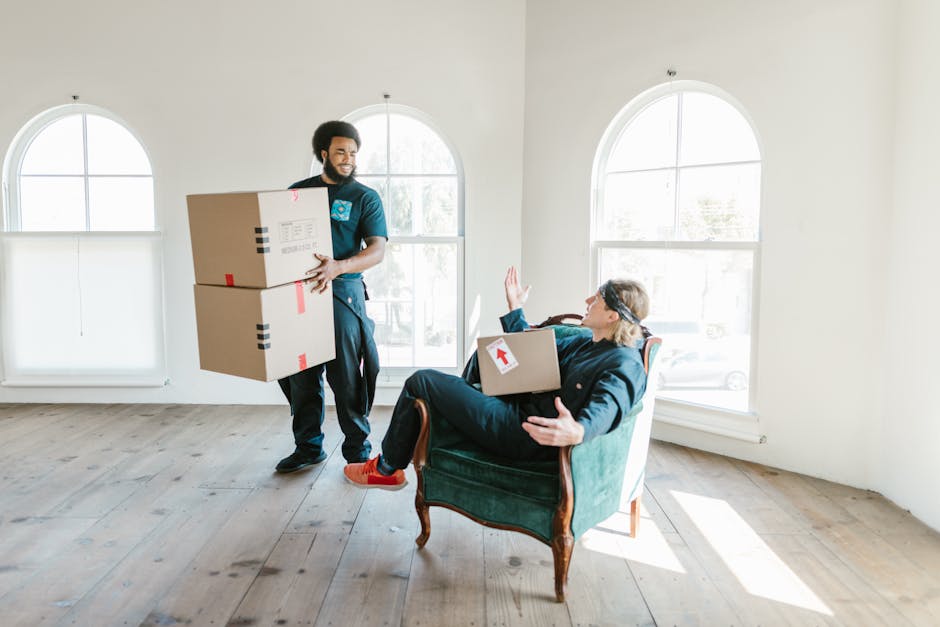 A man in a blue uniform and black pants is inside a bright, spacious room with light-colored wooden flooring and large, curved windows allowing natural daylight to illuminate the space. He is holding two medium-sized cardboard boxes taped securely with red and black packing tape, indicating they contain household items for a home relocation. Opposite him, a woman seated in a vintage-style armchair with wooden legs and green upholstery is raising her right hand in a gesture of greeting or farewell. She is wearing dark clothing and red shoes, with a cardboard box resting on her lap, marked with an upward arrow label for clear handling instructions. The scene captures the packing and moving process, with the man preparing to load the boxes onto a moving vehicle managed by Man and Van Highbury, specialists in removals and furniture transport. The environment suggests an organized packing session, with an emphasis on careful handling and transport logistics within a residential setting.