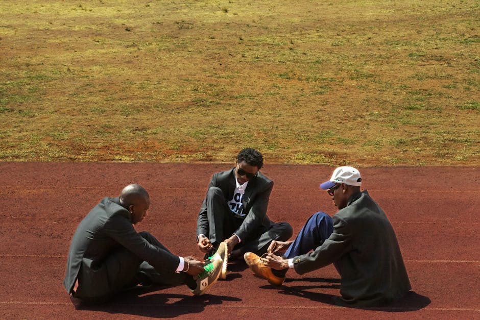 Inside a sports facility, three men are seated on a red running track with visible lane markings, engaging in a moment of preparation. Two of the men, dressed in dark suits and wearing sunglasses, are sitting cross-legged, while the third man, also in formal attire, is assisting one of the suited men by tying or adjusting his athletic shoes. The footwear is casual, with one pair featuring bright green laces. The background shows an area of grass or turf beyond the track, with natural lighting illuminating the scene. The setting suggests a break in activity or a moment of camaraderie during a sporting event or training session, which could be associated with the context of packing, moving, or logistical activities like those handled by Man and Van Highbury, though the action focuses on footwear and personal preparation rather than moving objects or furniture.