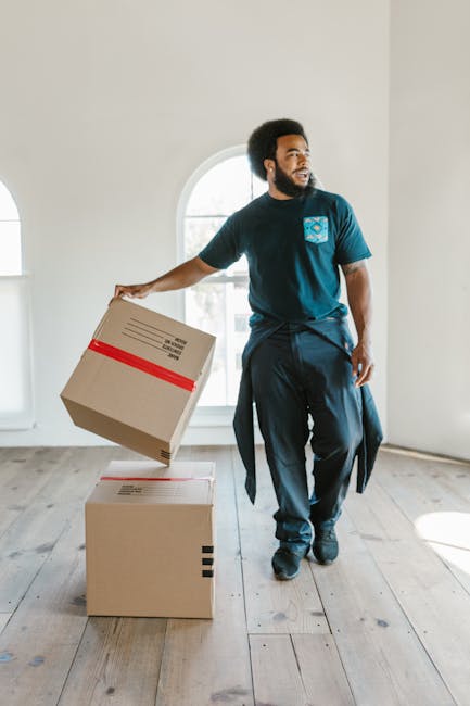 A man with dark curly hair and a beard, dressed in a navy blue t-shirt with a logo on the chest and dark trousers, is inside a bright, empty room with light wooden flooring and white walls. He is holding a medium-sized cardboard box with red tape in his right hand, preparing to place it on top of a larger, already stacked box on the floor. Behind him, there is a large arched window allowing natural light to fill the space. The scene appears to depict the process of home relocation, possibly as part of a professional removals service such as Man and Van Highbury, with the emphasis on packing and loading furniture and boxes for transportation. The environment suggests a professional, organized approach to moving, with no furniture or appliances visible, highlighting the packing and handling phase of a house move.