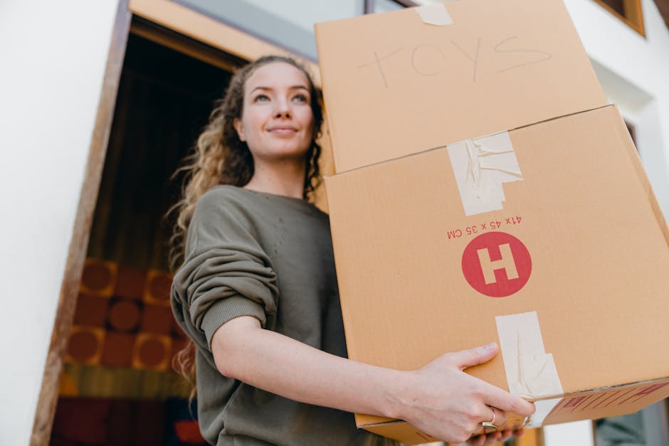 A woman with long, curly hair and a subtle smile is seen holding two large cardboard boxes filled with household items, likely during a house move. She is standing near a doorway inside a residential property, with a partially open door behind her revealing wooden interior elements. The boxes are sealed with packing tape, one labeled 'toys' written in marker, indicating contents of personal belongings for home relocation. Visible packing materials include cardboard boxes and packing tape. The woman’s clothing features a long-sleeved, loose-fitting top in a neutral tone, and her stance suggests she is in the process of loading or unloading these boxes as part of a furniture transport or packing and moving process. The environment is well-lit with natural light, emphasizing the practical aspect of the move supported by [COMPANY_NAME]'s house removals services.