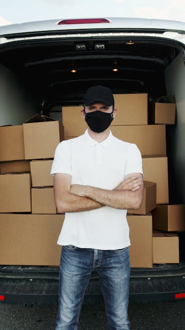 A mover dressed in a white protective suit, gloves, and a face mask is seen standing on a red brick driveway next to a white van loaded with brown cardboard boxes, some inside the vehicle and others n
